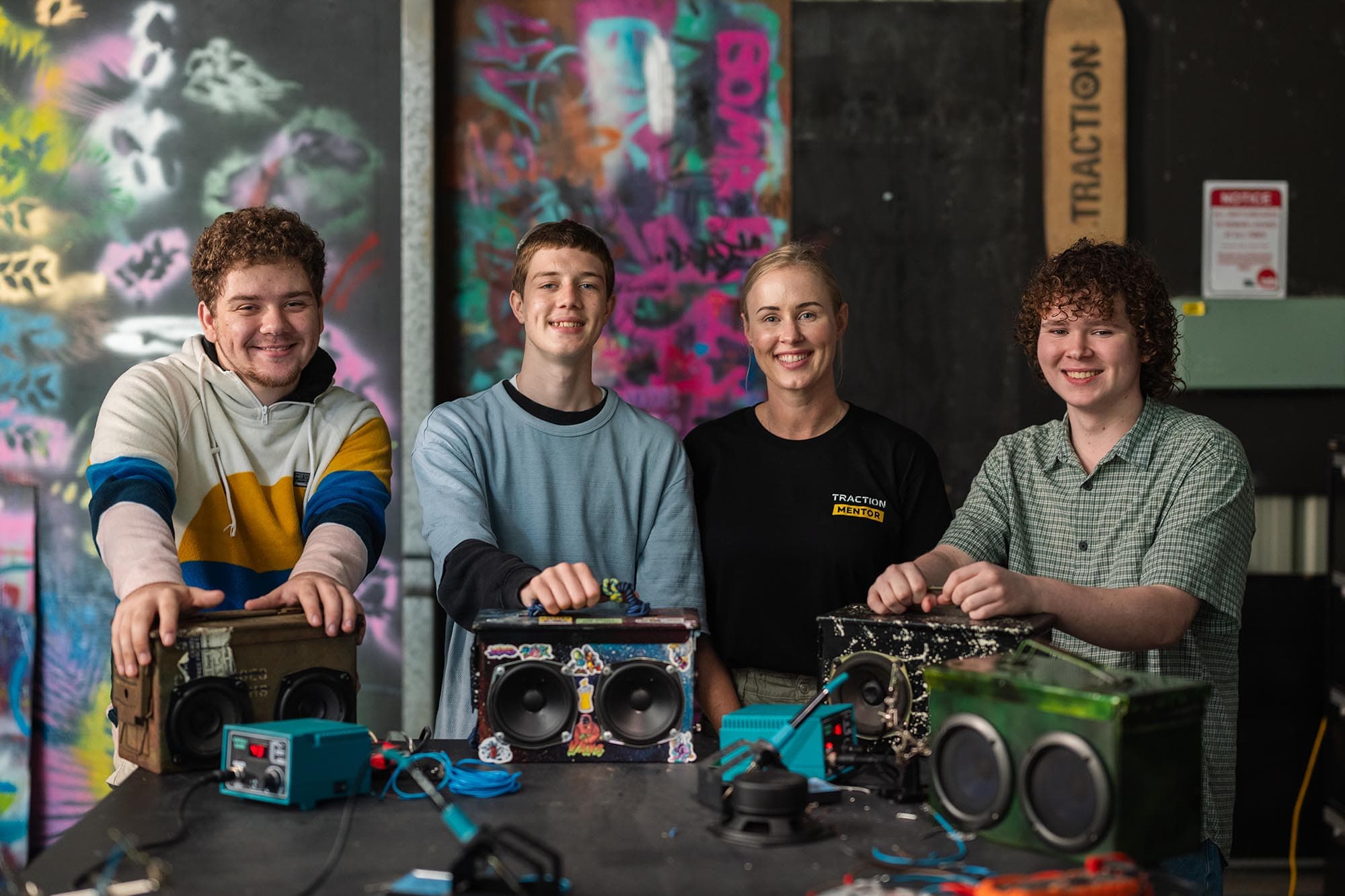 A photo of four people—three young men and one woman—standing behind a table in a workshop setting. They are all smiling at the camera, with their hands resting on custom-made, portable boomboxes they appear to have built. The woman is wearing a black shirt that says "TRACTION MENTOR." The background is dark and covered in colorful graffiti.