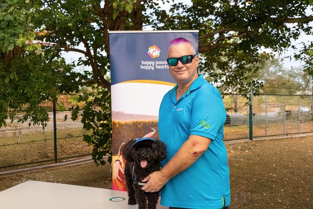 A man with bright purple hair and sunglasses, identified as David, stands next to a banner for "Happy Paws Happy Hearts." He is wearing a blue polo shirt and is holding a small black dog that has a blue baseball cap on its head. The man is smiling at the camera.