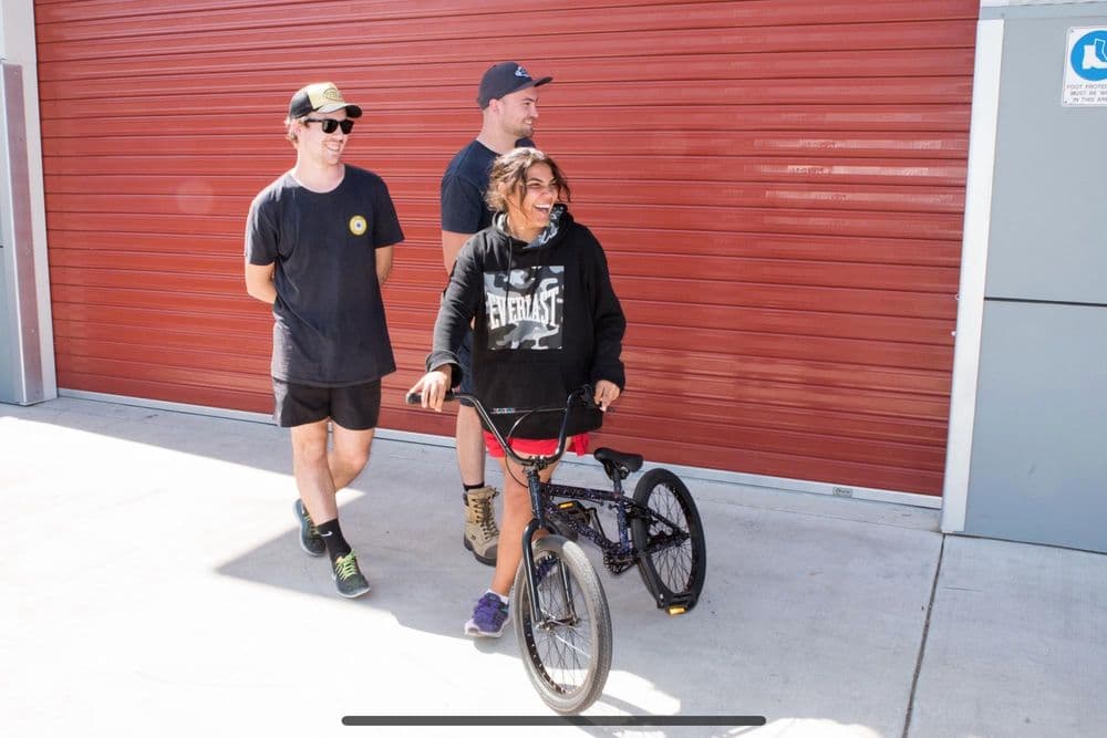 A young woman, Leila, wearing a black Everlast hoodie and red shorts, stands next to her BMX bike, which has a custom two-wheel attachment on the back. She is smiling and looking off to the side. Two young men are standing behind her in front of a large red roller door.