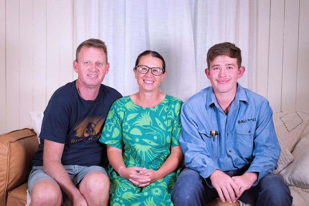 A portrait of three people—two men and one woman—sitting together on a brown leather couch in front of a white curtain. The woman in the center wears a bright green patterned dress and glasses. The young man on the right, Louis, wears a blue work shirt, and the older man on the left wears a navy t-shirt and shorts. They are all smiling at the camera.