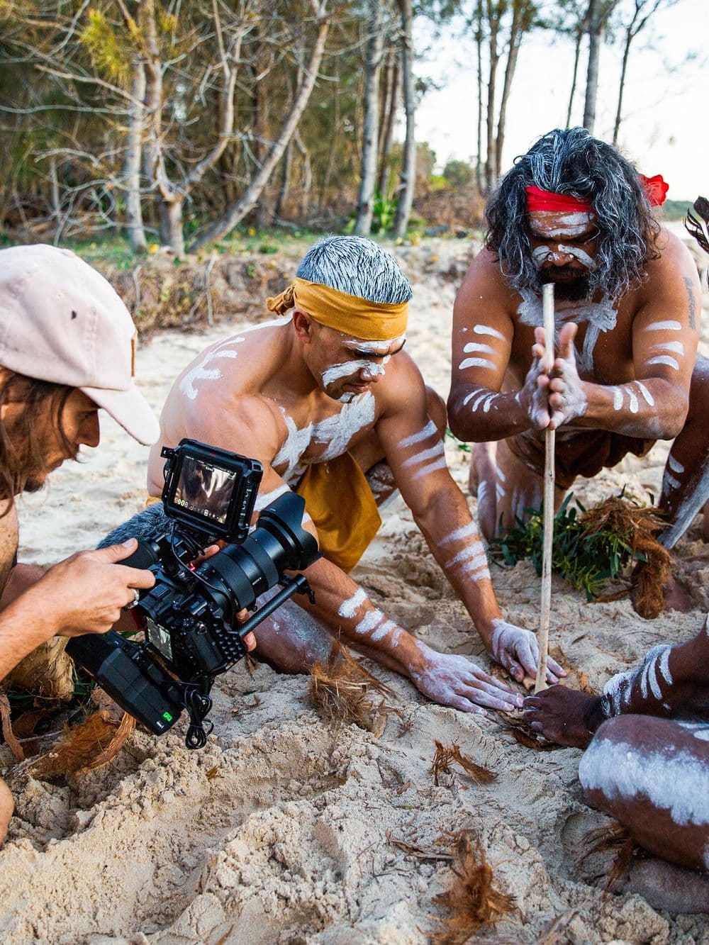 A person with a camera is on a sandy beach, filming two Indigenous men. The men are wearing traditional body paint, and one of them is using a stick to make fire by rubbing it against the ground. They are surrounded by trees.