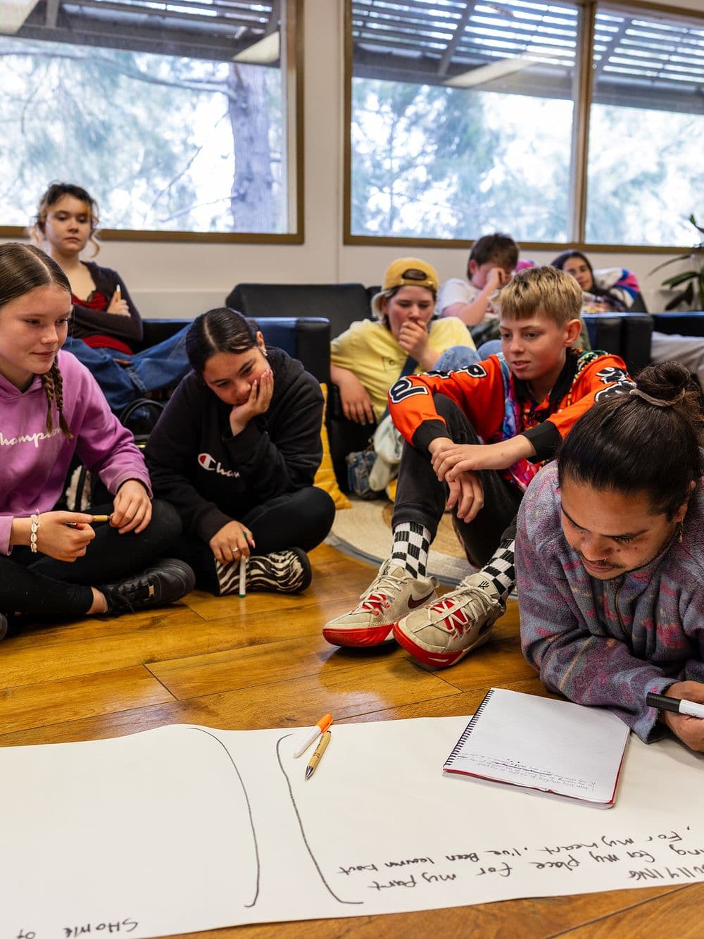 A group of young Indigenous people sit on a wooden floor around a large sheet of paper. Several of them are writing, drawing, and talking. The scene appears to be an indoor workshop or meeting.