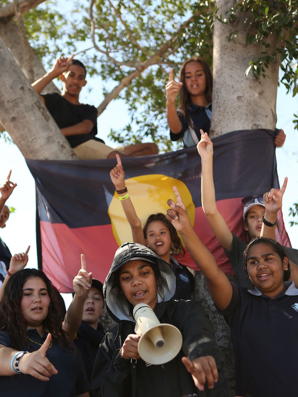 A large group of young Indigenous people are gathered under a tree, holding a red, yellow, and black Aboriginal flag behind them. Several people are raising their fists, and a young woman in a black hoodie is speaking into a megaphone.