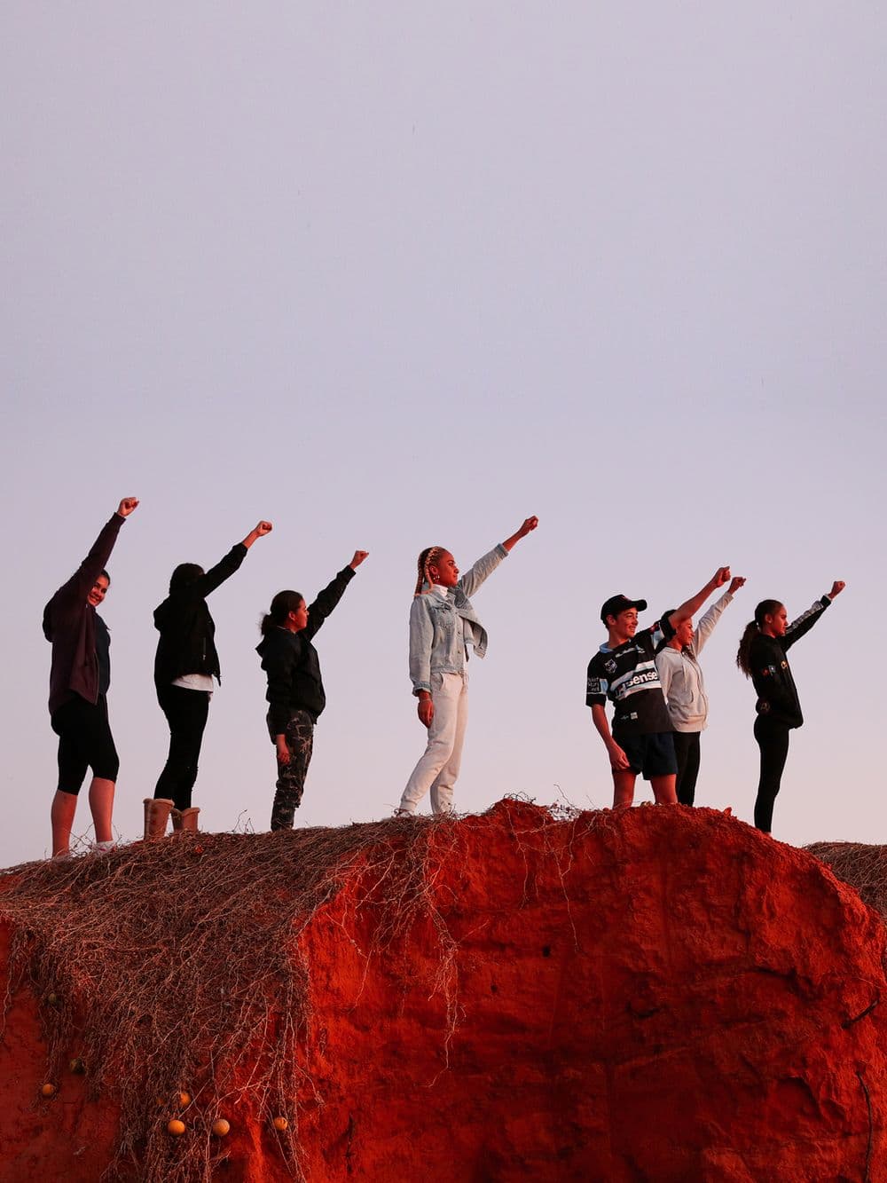 A group of seven young people are silhouetted against a bright sky, standing on a red dirt mound. Each person has one arm raised and pointed forward, as if they are superheroes.