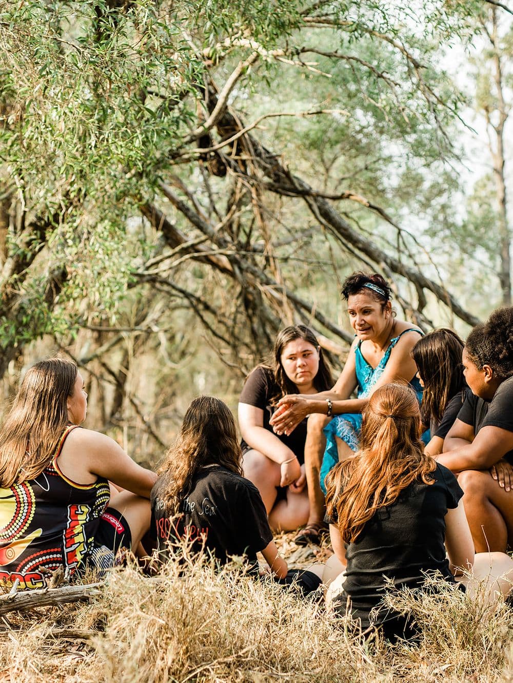 A group of five young Indigenous people are sitting on the ground in a circle with an adult woman in a blue top. They are in a natural setting with tall grass and trees. The woman is gesturing and talking to the group, which appears to be in an outdoor discussion circle.