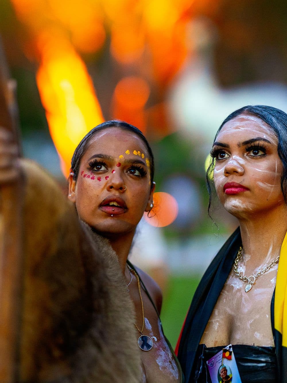 A close-up portrait of two young Indigenous women with traditional body paint on their faces and chests. They are looking up and to the side with serious expressions. The woman on the left has yellow dots on her face, and the woman on the right has white paint. A fire is blurred in the background.