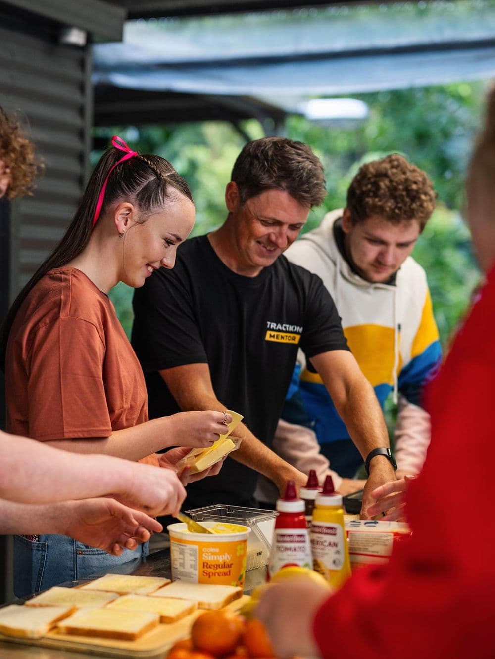 A group of young people and a male mentor, who is wearing a black "Traction Mentor" shirt, are gathered outdoors making sandwiches. They are standing around a table with bread, butter, and condiments. The mentor is smiling down at the food he is preparing.